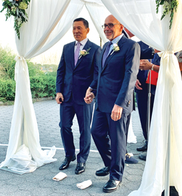 Two men clasp hands and smile under a white chuppah.